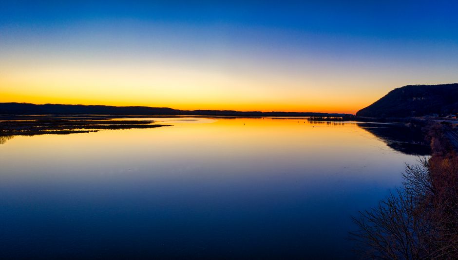An expansive, serene view of a wide river reflecting the warm, golden light of a stunning sunset, with rolling hills in the background.