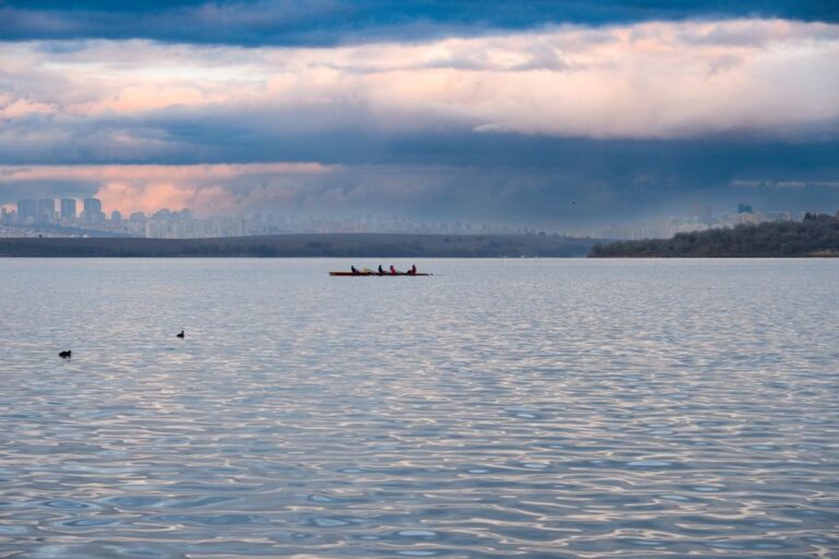 Lone rower in a scull pauses on calm, glassy water during an atmospheric sunrise, conveying a sense of introspection and a...